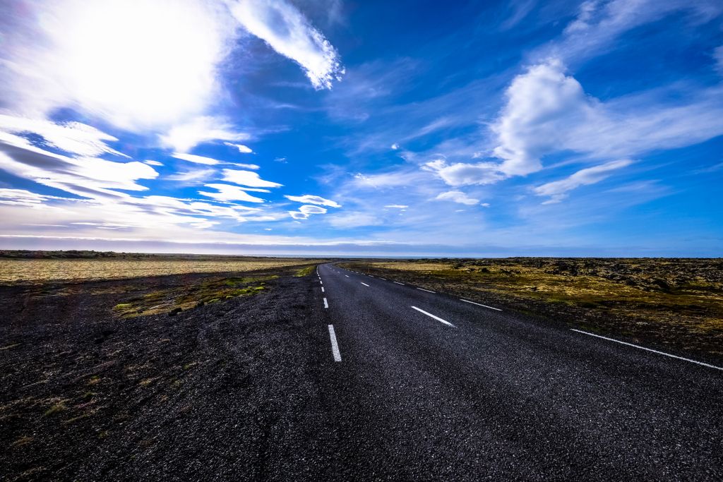 Long empty highway stretching under a vivid blue sky with scattered clouds in a rural landscape.
